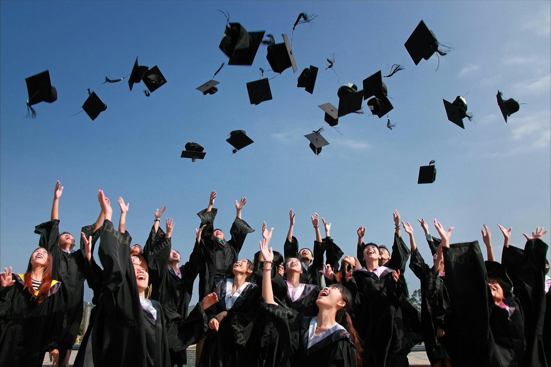 newly graduated college students wearing black academy gowns throwing hats up in the air
