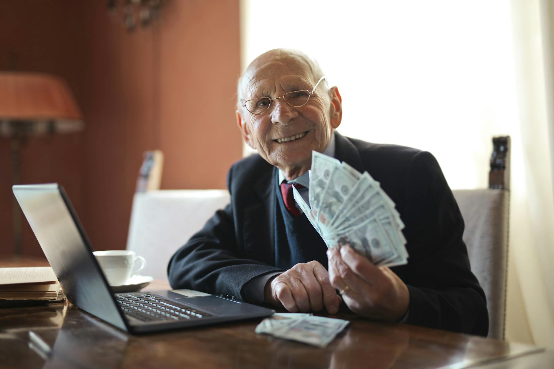 happy senior businessman holding money in hand while working on laptop at table How to Protect Your Money: 5 Habits for Long-Term Wealth