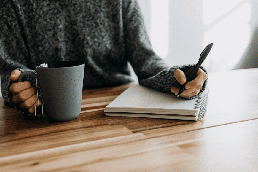 10 Steps to Build a Successful Writing Business 7 A person wearing a cozy sweater holding a gray mug in one hand and writing in a notebook with a pen on a wooden table. 10 Steps to Build a Successful Writing Business