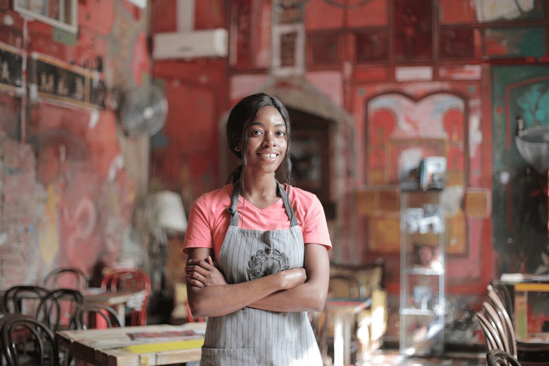 How to Make Clients Feel Valued Before You Even Speak 7 A woman wearing an apron stands confidently with her arms crossed in a colorful, artistic cafe interior. How to Make Clients Feel Valued Before You Even Speak