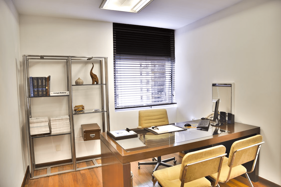 A modern office setting featuring a wooden desk with a glass top, an ergonomic chair, and a shelving unit displaying books and decorative items. The room has ample natural light from a window with blinds. 10 Ways to Make Clients Feel Comfortable in Your Office