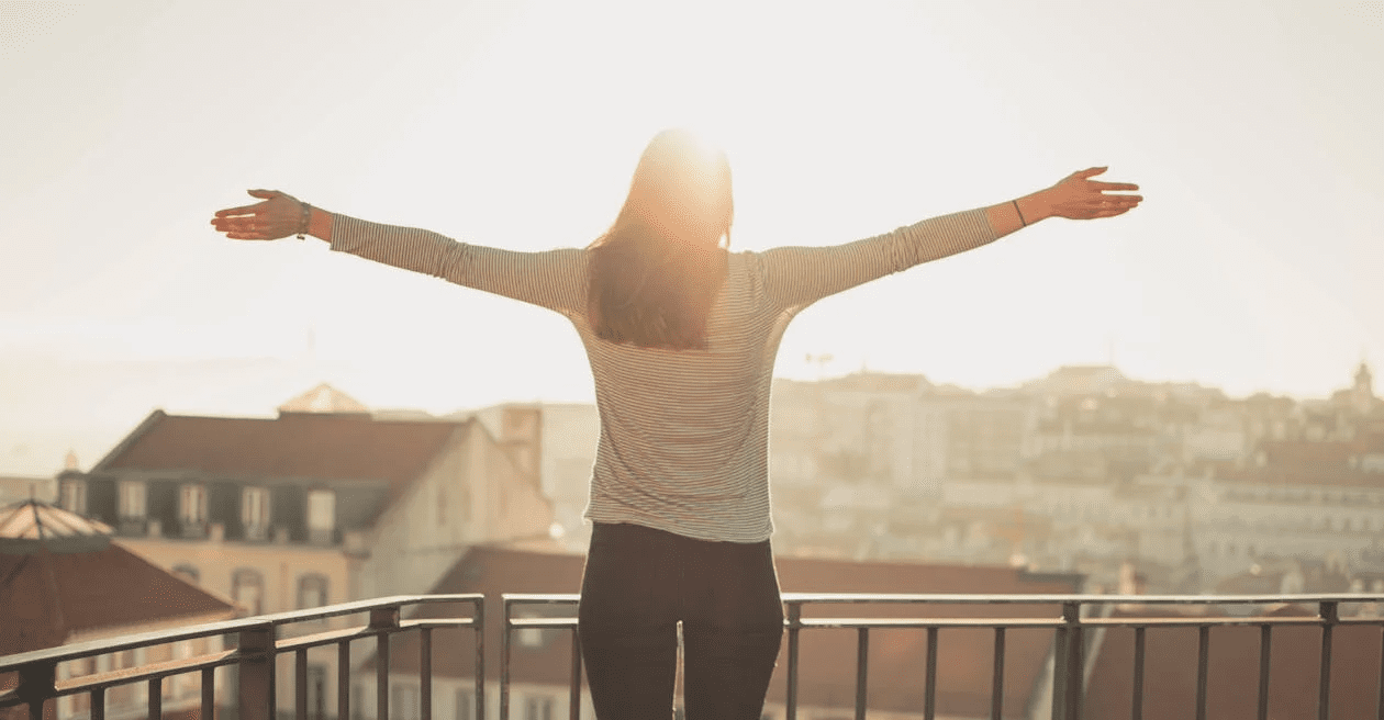 6 Empowering Ways to Reclaim How You Feel About Yourself 13 A woman with her arms outstretched, standing on a balcony with a city view in the background, illuminated by sunlight. 6 Empowering Ways to Reclaim How You Feel About Yourself