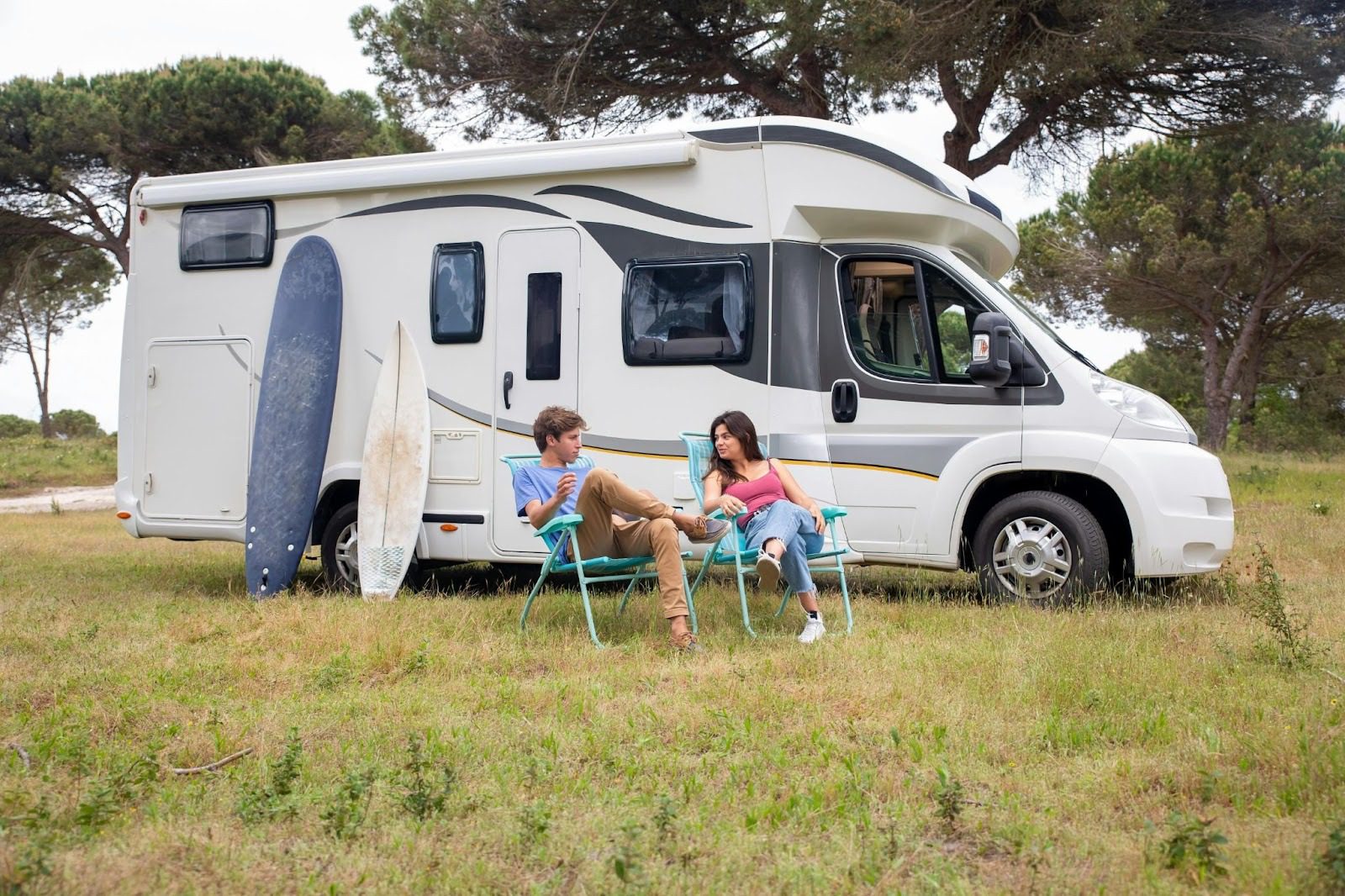 A couple sitting on folding chairs beside a parked RV, with surfboards leaning against the vehicle, in a grassy field surrounded by trees. Road Trip Tips for Comfort and Adventure