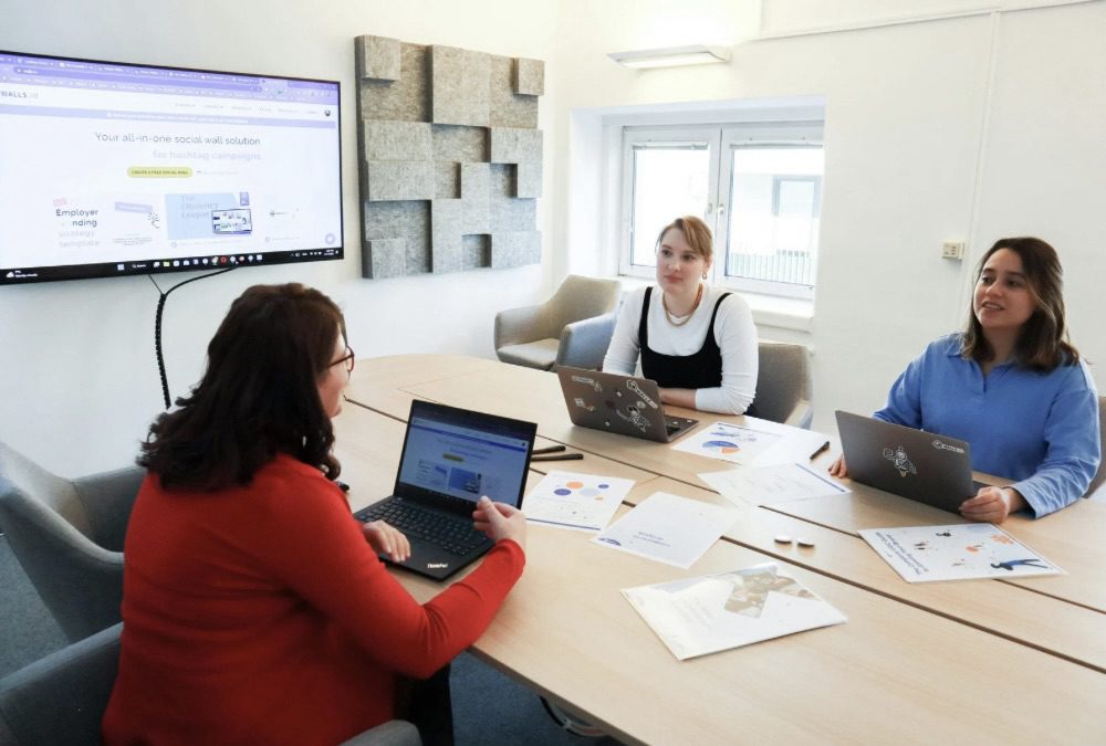 Three people in a meeting room discussing a marketing strategy, with laptops open and a screen displaying a social media solution. 10 Proven Tips for Finding Help with Your Marketing Strategy