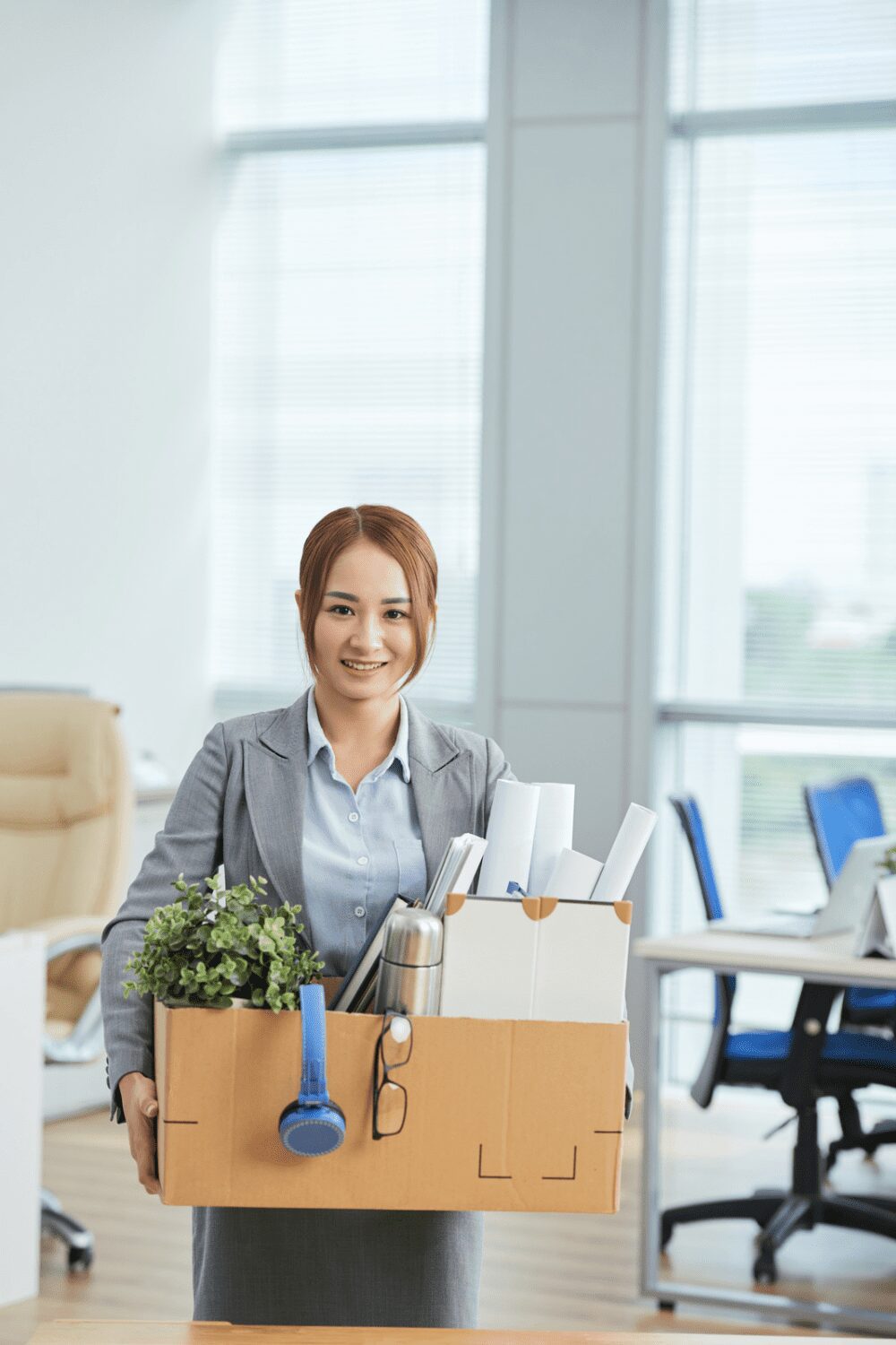 Relocating for Work: 6 Smart Strategies for a Smooth Move 13 A woman in a business suit is smiling while carrying a cardboard box filled with office supplies, a small plant, a thermos, and some documents in a modern office setting. Relocating for Work: 6 Smart Strategies for a Smooth Move