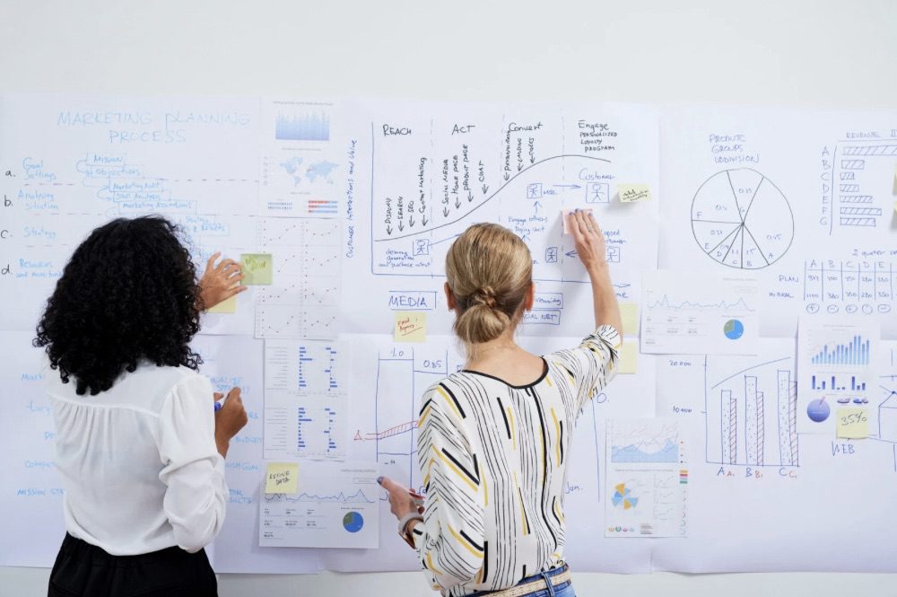 Two individuals collaborating on a marketing strategy, analyzing graphs and charts displayed on a wall, with sticky notes and documents surrounding them. 10 Proven Tips for Finding Help with Your Marketing Strategy