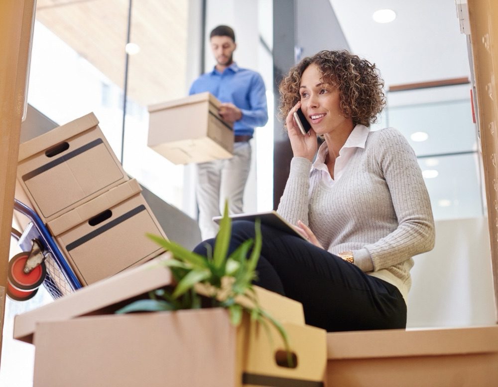 A woman sitting on stairs surrounded by moving boxes, talking on the phone while a man carrying a box walks in the background. Relocating for Work: 6 Smart Strategies for a Smooth Move