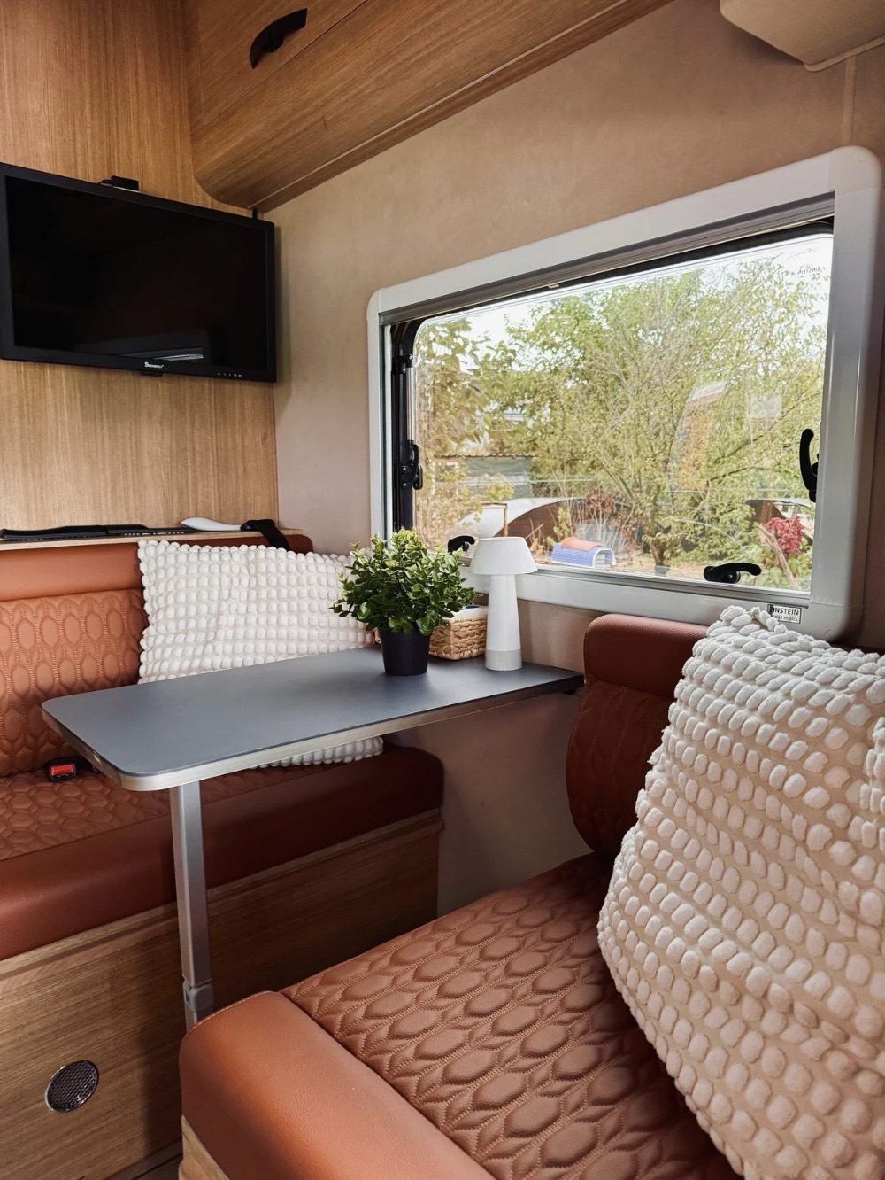 Cozy interior of an RV featuring a dining area with a gray table, plush cushions, a small lamp, and a view of greenery visible through a window. Road Trip Tips for Comfort and Adventure