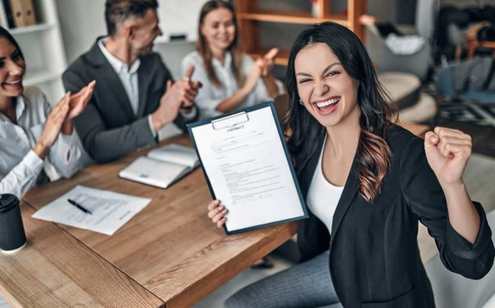 A woman celebrating her job offer with a contract in hand, surrounded by supportive colleagues clapping in an office setting. Relocating for Work: 6 Smart Strategies for a Smooth Move