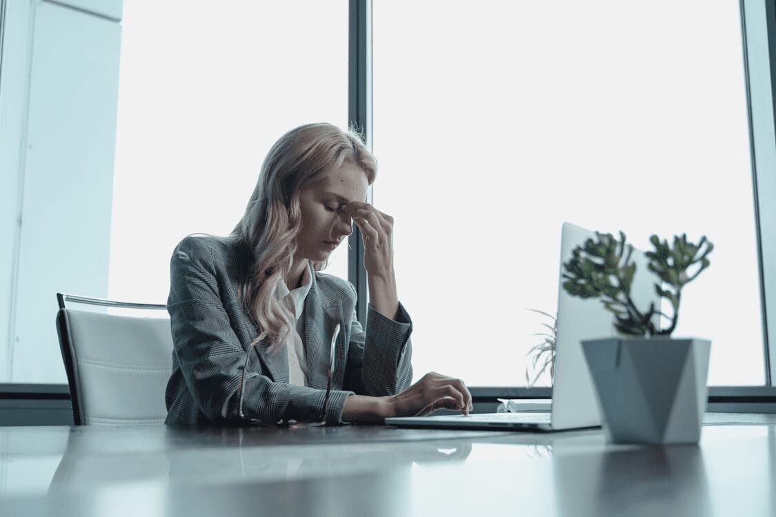 How Bad Marketing Can Hurt More Than Just Your Business 7 A woman in a blazer sitting at a desk with a laptop, appearing stressed while rubbing her forehead, in a modern office setting with large windows. How Bad Marketing Can Hurt More Than Just Your Business