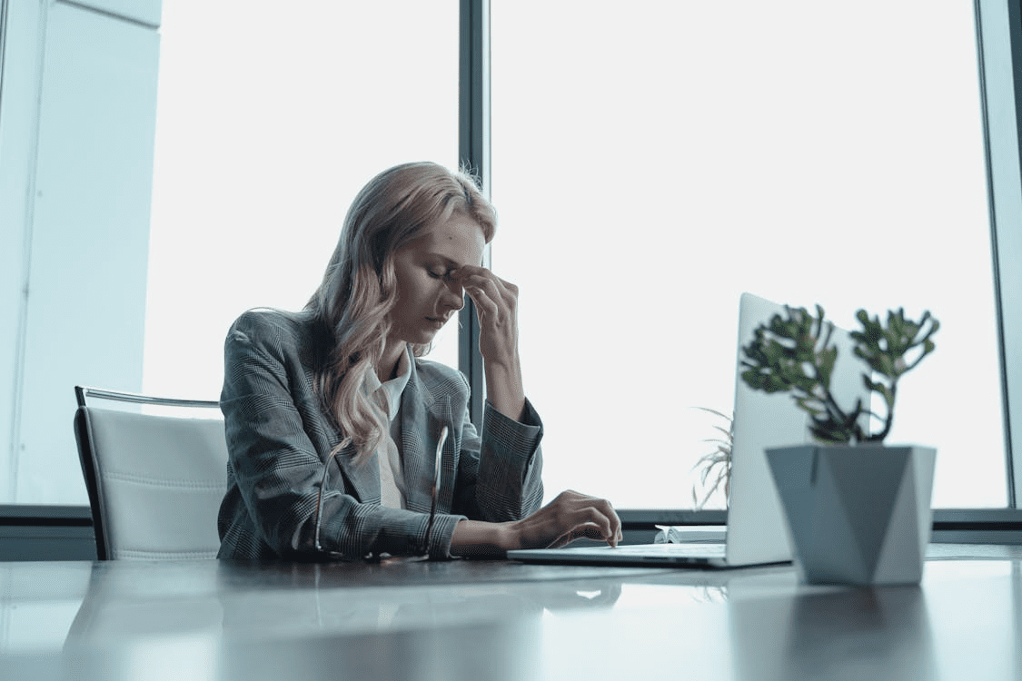 A woman in a blazer sitting at a desk with a laptop, appearing stressed while rubbing her forehead, in a modern office setting with large windows. How Bad Marketing Can Hurt More Than Just Your Business
