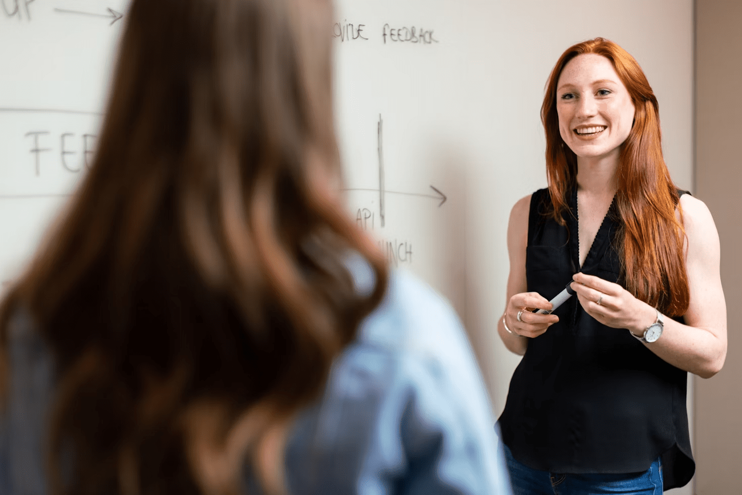 6 Careers That Let You Travel and Work At The Same Time 11 A woman with long red hair and a black sleeveless top smiles and engages with another person in a professional setting, as she holds a marker and stands in front of a whiteboard. 6 Careers That Let You Travel and Work At The Same Time
