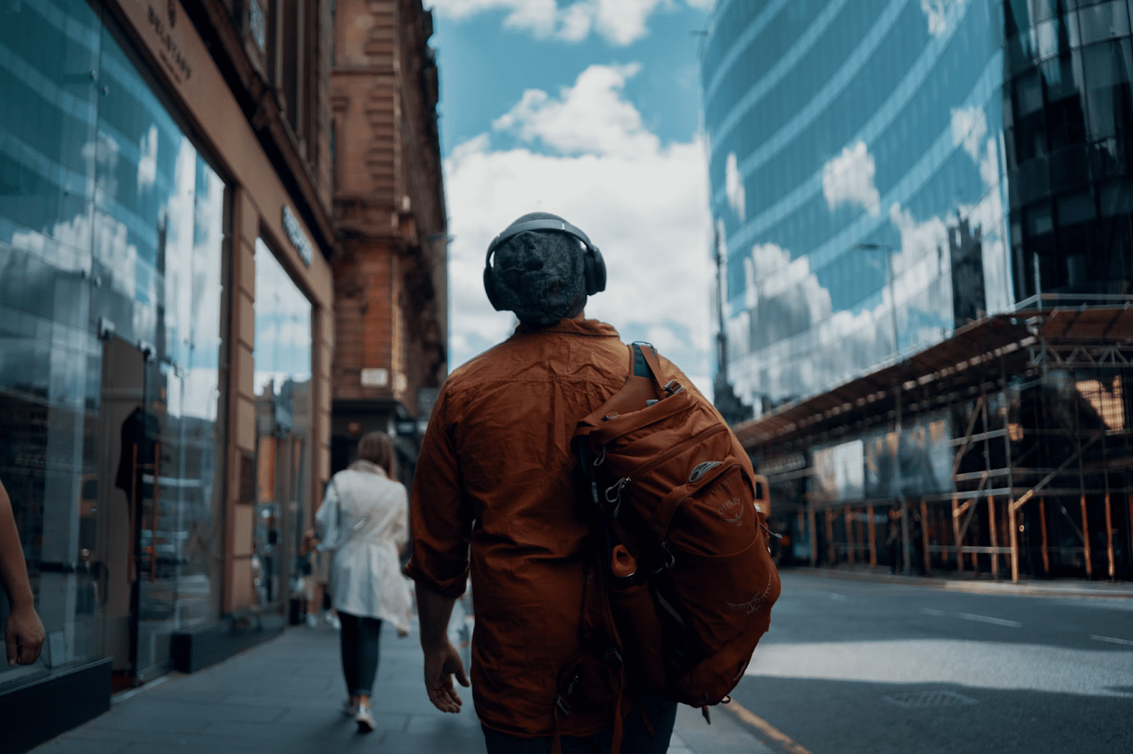 Person wearing headphones and an orange jacket walking on a city street with modern buildings in the background. Sustainability Mindset: 7 Ethical Ways to Cut Carbon on Your Commute