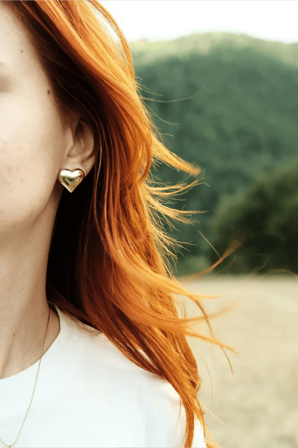 A close-up of a woman with vibrant red hair wearing heart-shaped gold earrings, with a blurred natural landscape in the background. Best Diamond Gold Stud Earrings for Women