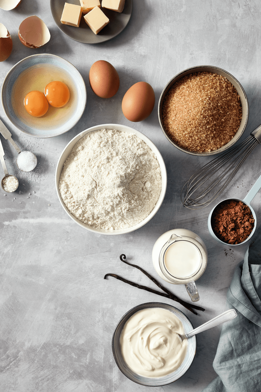An assortment of baking ingredients including eggs, flour, brown sugar, butter, milk, cocoa powder, and vanilla on a gray surface, arranged with measuring spoons and a whisk. 30 Essential Winter Pantry Staples for a Self-Sufficient Kitchen