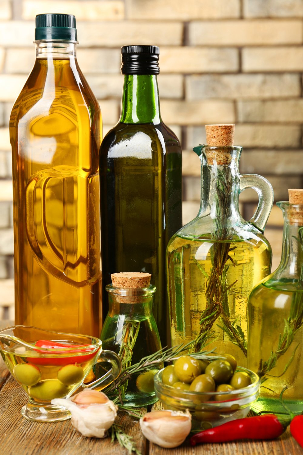 An assortment of various oils in glass and plastic bottles, alongside garlic cloves, green olives, and chili peppers, arranged on a wooden surface. 30 Essential Winter Pantry Staples for a Self-Sufficient Kitchen
